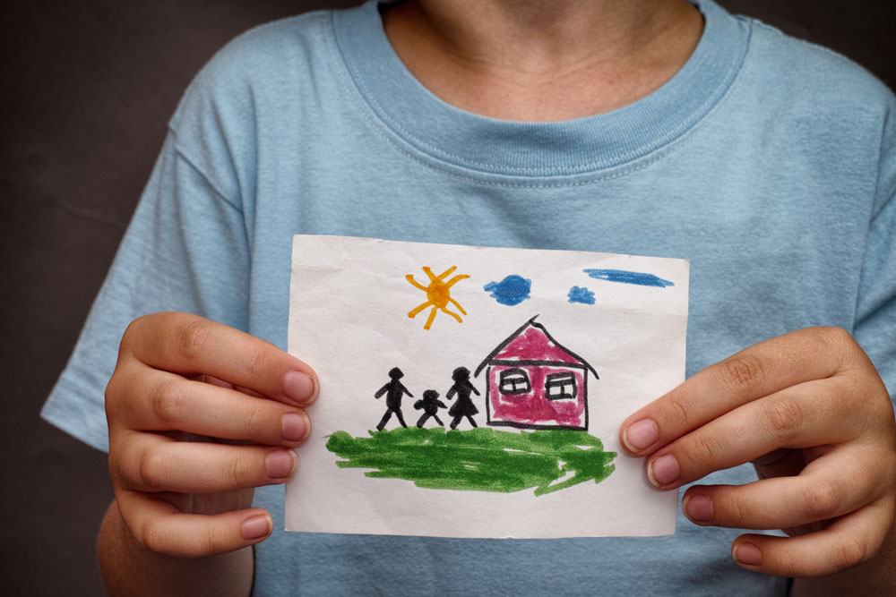 Child holding a drawing of a house and family playing outside.