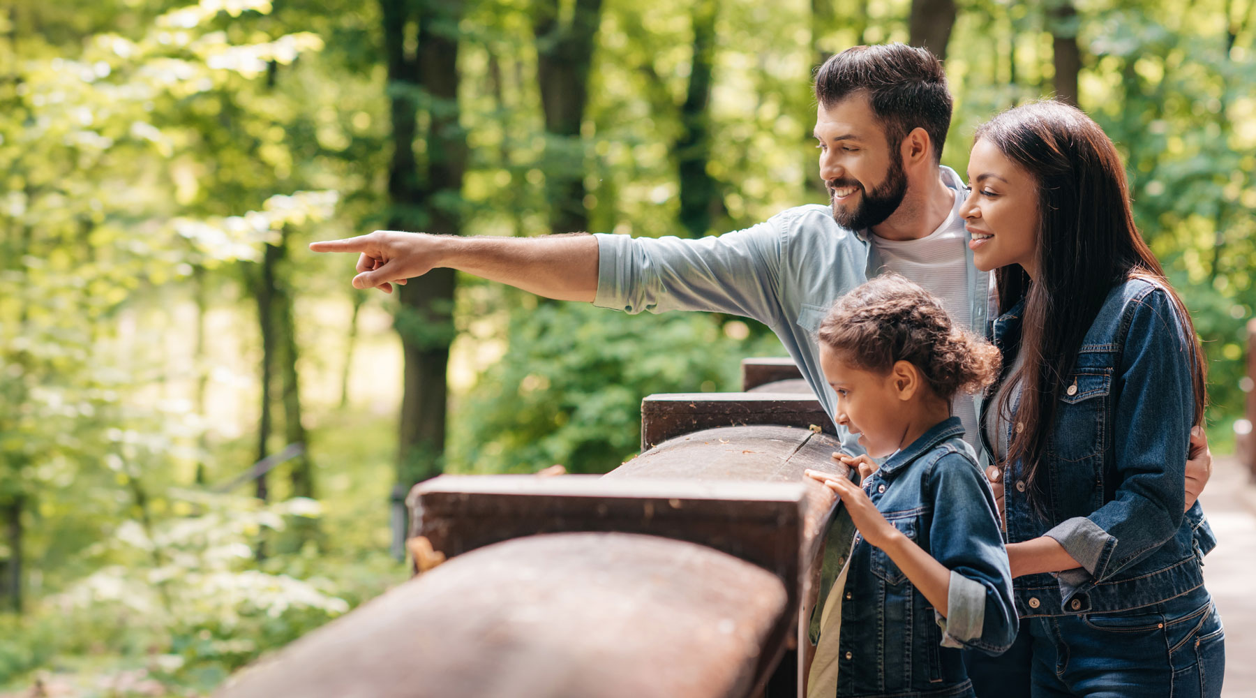Young smiling interracial family standing on wooden bridge, while father pointing somewhere into the forest