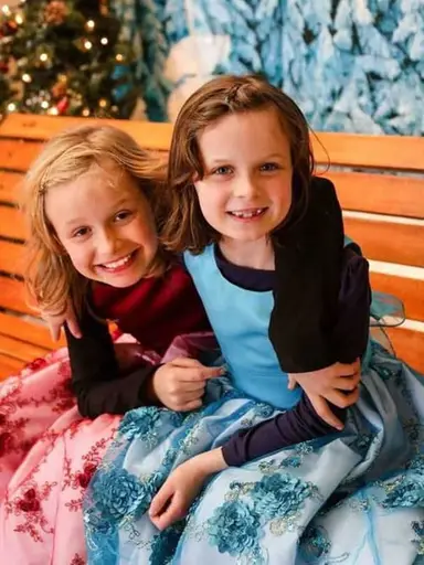 Two young girls sitting on a bench wearing dresses.